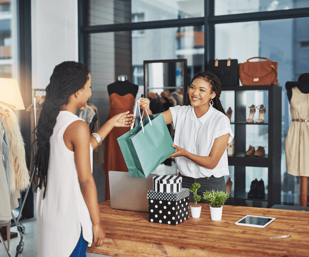 Customer receiving a shopping bag from a store employee at a fashion boutique with clothing and accessories displayed.