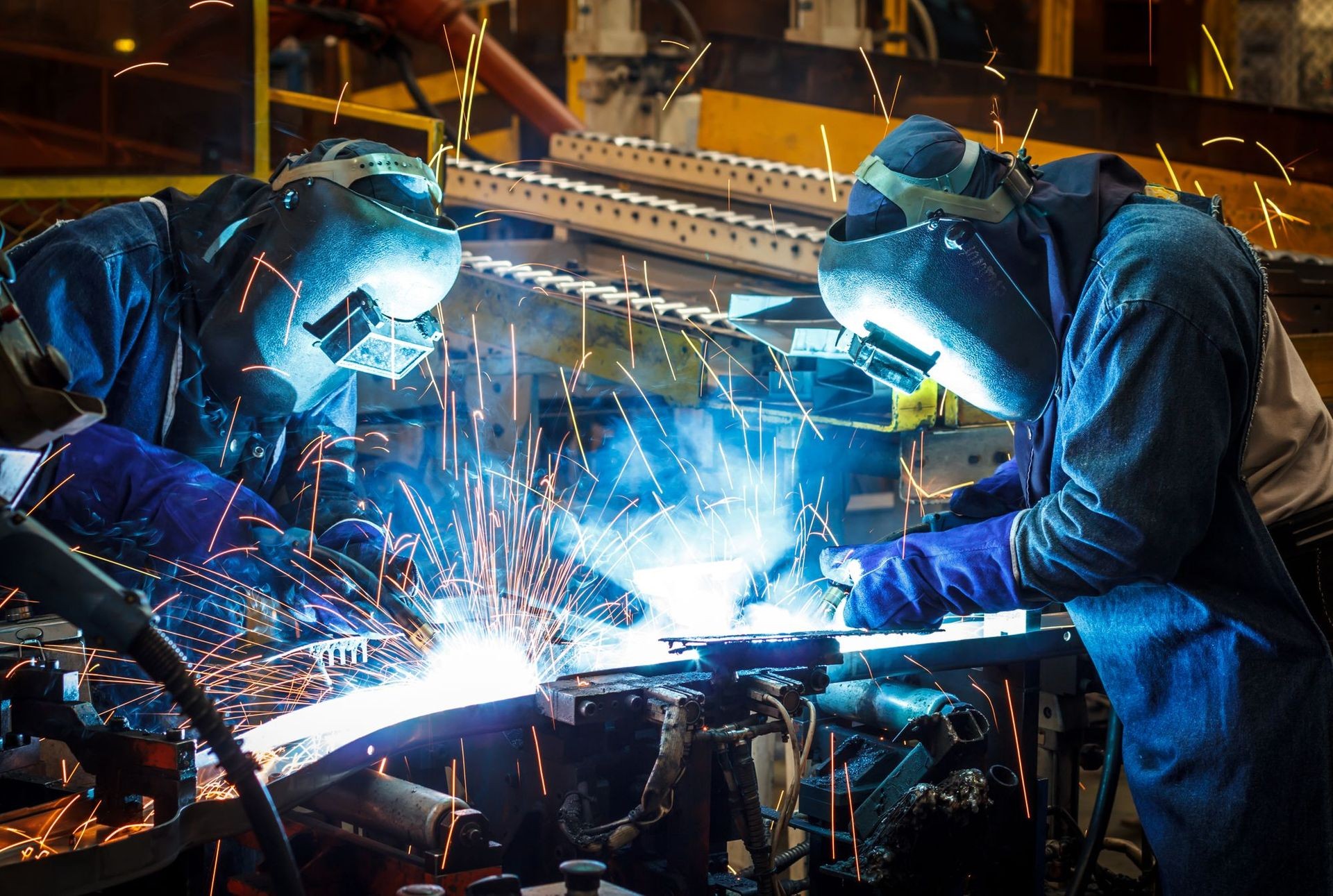 Two industrial workers wearing protective gear perform welding in a factory with sparks flying from the bright weld.